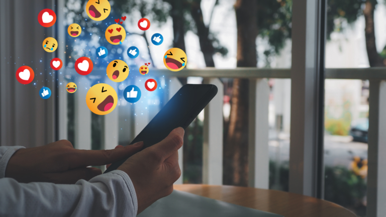 Hands holding a smartphone with social media reaction emojis like likes, hearts, and laughter floating above the screen.