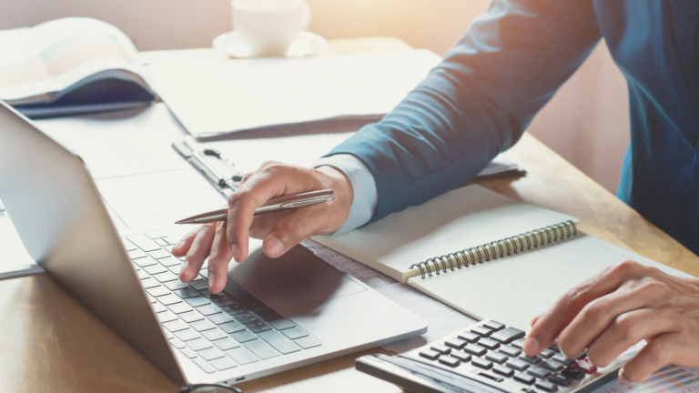 Professional working on a laptop and calculator at a desk, analyzing finances and data with a notepad nearby.